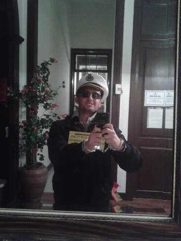 Un homme habillé en capitaine, portant des lunettes de soleil et un chapeau blanc, prend un selfie miroir à l'intérieur. Il y a une plante et des portes en bois à l'arrière-plan.