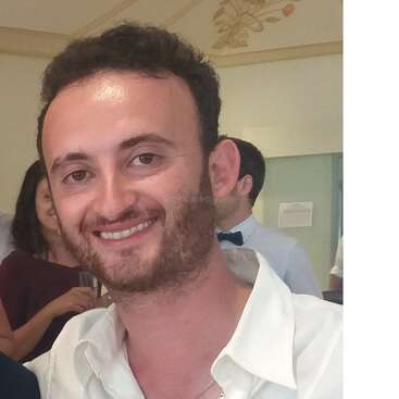 The image shows a man with short dark hair and a white shirt, smiling at the camera, with other people in the background.