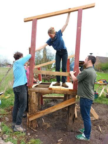 Tres personas trabajan juntas al aire libre montando una estructura de madera. Una de ellas está de pie sobre la plataforma, equilibrando una viga, mientras que las otras sujetan y sostienen el armazón en su sitio.