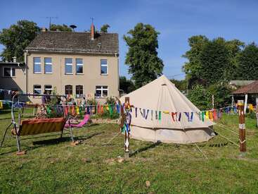 En un jardín cubierto de césped, decorado con banderines de colores, hay una gran carpa de color beige. Cerca hay un columpio y un tobogán, y al fondo se ve una casita.