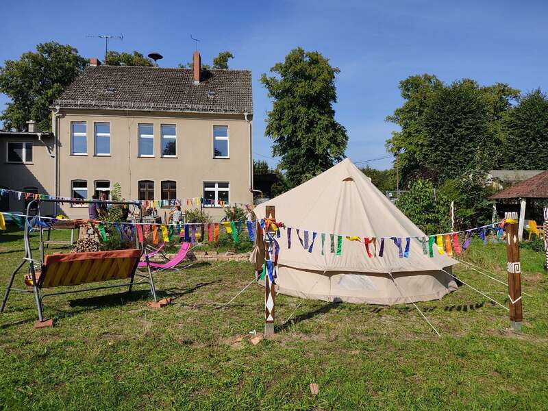 En un jardín cubierto de césped, decorado con banderines de colores, hay una gran carpa de color beige. Cerca hay un columpio y un tobogán, y al fondo se ve una casita.
