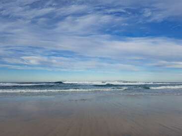 A tranquil beach scene with gentle waves rolling onto wet sand. The sky is expansive, filled with soft, wispy clouds and shades of blue above the horizon.