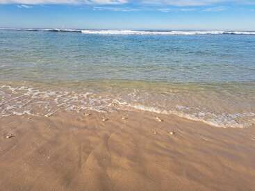 Golden sand meets gentle, clear blue waves under a bright sky. White clouds are scattered above, creating a peaceful and serene beach atmosphere.