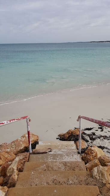 The image depicts a serene beach scene with a staircase leading down to the shore, flanked by rocks and featuring red and white handrails.