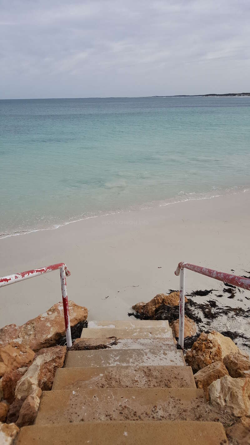 The image depicts a serene beach scene with a staircase leading down to the shore, flanked by rocks and featuring red and white handrails.
