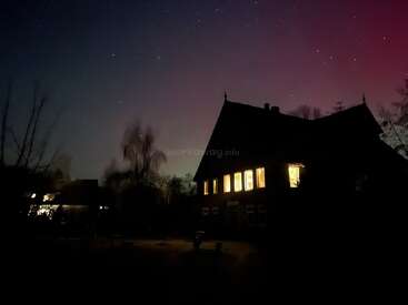Una casa oscura con ventanas iluminadas se alza bajo un cielo estrellado. Árboles recortados contra el cielo la rodean, y un sutil tono púrpura tiñe el horizonte.