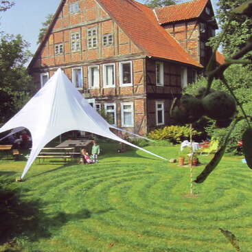 L'image représente une grande maison à plusieurs étages avec un toit rouge et une tente blanche au premier plan, entourée d'une pelouse verdoyante et d'arbres.