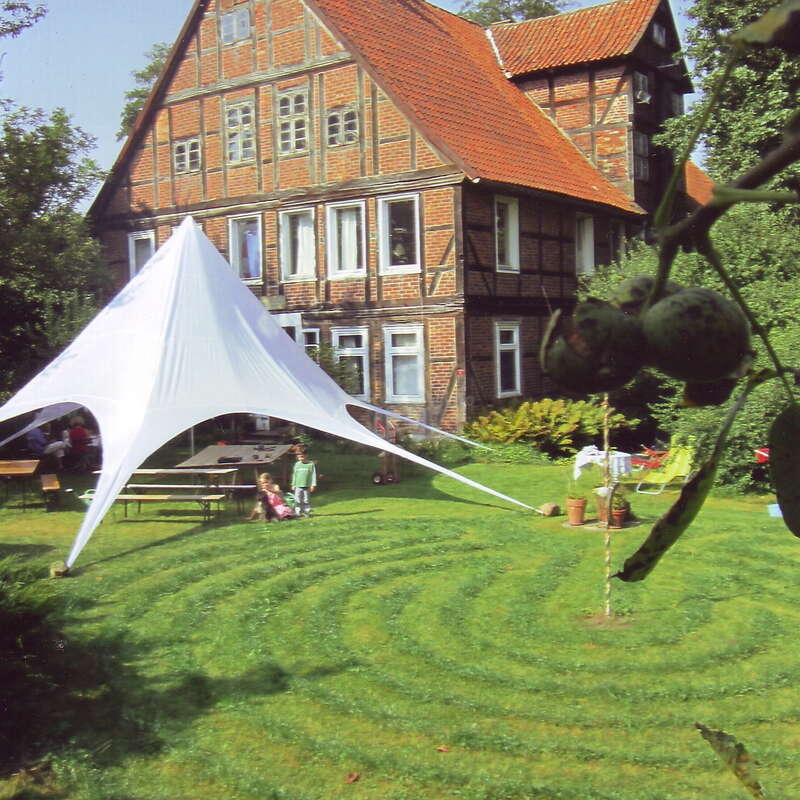 The image depicts a large, multi-story house with a red roof and a white tent in the foreground, surrounded by a lush green lawn and trees.