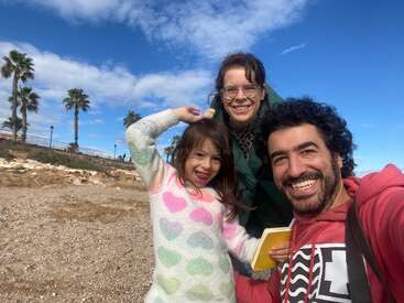 Eine fröhliche dreiköpfige Familie posiert am Strand unter Palmen vor blauem Himmel. Das kleine Mädchen trägt einen Pullover mit Herzmuster, und alle strahlen vor Freude.