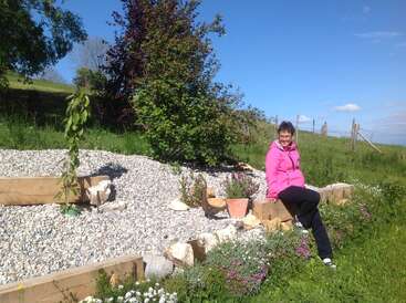 A woman in a bright pink jacket sits on a wooden border in a sunny garden with gravel, potted plants, green grass, trees, and blue sky.