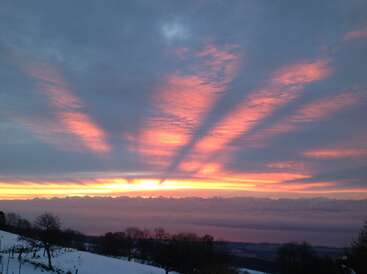 Radiant sunrise with dramatic crepuscular rays fan across a colorful sky. Snow-covered landscape and bare trees enhance the serene, breathtaking beauty of nature’s early morning.