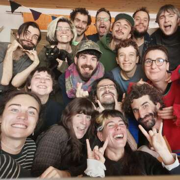 A group of joyful people poses together indoors, making silly faces and hand gestures. They look happy, energetic, and united. A black cat is included, too.