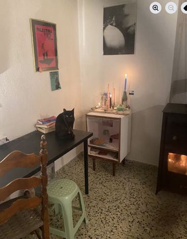 Une pièce confortable et faiblement éclairée contient un chat noir sur un bureau, des livres, des affiches, des bougies, un petit meuble, une chaise en bois et un poêle allumé.