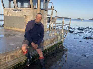 Un homme est assis sur le bord d'un bateau nommé « Emmanon », vêtu de bottes en caoutchouc et d'un pull, avec une mer calme et des îles lointaines derrière lui.