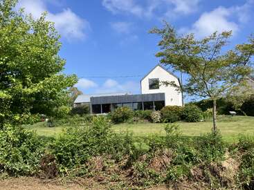 Une maison moderne avec une façade blanche et un toit gris se trouve au milieu d'un jardin verdoyant. Les arbres, les buissons et le ciel bleu créent une atmosphère paisible et accueillante.