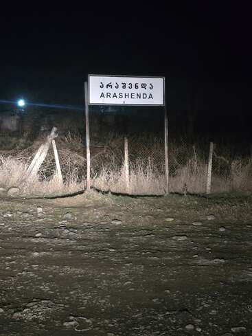 Un poste indicador se alza en la noche, iluminado por los faros, mostrando "ARASHENDA" en escritura inglesa y georgiana. A su alrededor, un camino de tierra áspera y hierba seca.