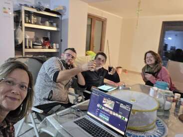 Cuatro personas se sientan alrededor de una mesa, sonrientes y conversando. Enfrente hay un portátil abierto y varios objetos esparcidos por la mesa. Ambiente acogedor.