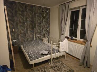 Un dormitorio acogedor con papel pintado estampado, cama de hierro y ropa de cama gris. Hay una ventana con cortinas, una alfombra y una lámpara colgante sobre la cama.