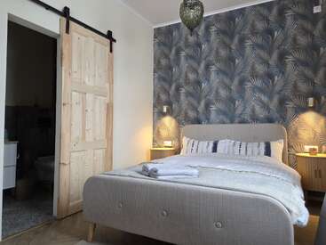 A cozy bedroom features a double bed with neatly arranged pillows, soft lighting, tropical wallpaper, a wooden sliding door, bedside tables, and folded towels on the bed.