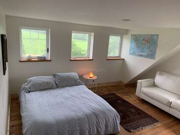 The image depicts a serene bedroom with a bed, couch, and rug, featuring three windows and a world map on the wall, exuding a cozy and peaceful atmosphere.