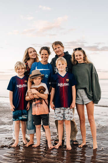 The image depicts a family of seven standing on a beach, with two adults and five children, all wearing casual clothing and smiling at the camera.