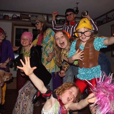 A group of people, mostly children, wear colorful, funky costumes and pose energetically indoors, creating a lively, playful, and joyful party atmosphere.