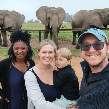 Quatre personnes souriantes, dont un jeune enfant, posent ensemble en plein air. Derrière eux, trois éléphants se tiennent sur un terrain herbeux, créant ainsi une expérience unique et joyeuse de la vie sauvage.