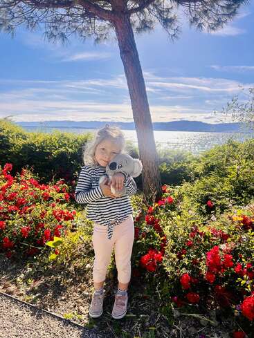 A young girl stands under a tree, holding a stuffed koala. She’s surrounded by vibrant red flowers, with a scenic lake and mountains behind her.
