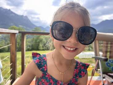 A smiling young girl wears oversized black sunglasses and a floral dress, sitting outdoors. Behind her are mountains, green fields, and a cloudy sky. Joyful summer moment.