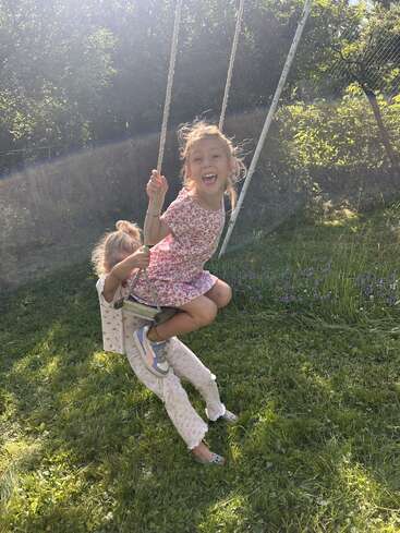 Two young girls share a swing on a sunny day, smiling and laughing in a lush green garden, surrounded by trees and wildflowers, enjoying childhood joy.