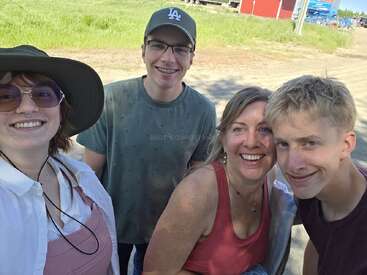Four people, smiling outdoors on a sunny day, stand close together for a selfie. They appear relaxed and happy, with green grass and blue sky behind them.