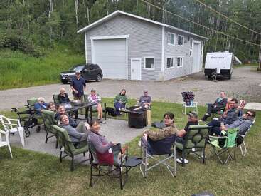 A group of people sits in a large circle outdoors, enjoying a gathering near a fire pit. There are lawn chairs, a garage, trees, and a camper.