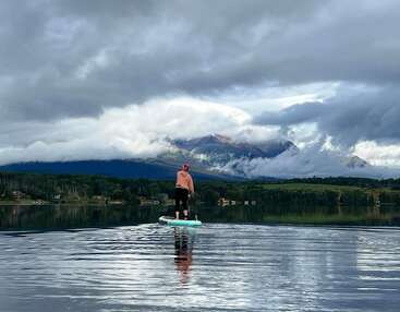 A person in a pink hoodie and black pants paddleboards on a calm lake, surrounded by lush forests and dramatic, cloud-covered mountains under a moody sky.
