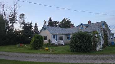 A cozy beige house with blue trim sits surrounded by lush green grass, bushes, and colorful flowers, backed by tall trees under a clear sky.