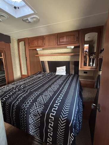Cette image montre une chambre confortable en camping-car avec un couvre-lit à motifs noirs et blancs, des armoires en bois au-dessus et à côté du lit, un placard à miroir et un sol recouvert de moquette.