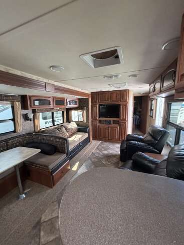 Cette image montre l'intérieur d'un véhicule récréatif avec une salle à manger confortable, des fauteuils inclinables en cuir, un canapé, de grandes fenêtres, des armoires en bois et une télévision.