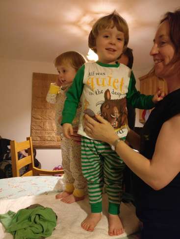 La imagen muestra a una mujer ayudando a un niño pequeño a ponerse un pijama de rayas verdes y blancas, con otro niño de pie cerca.