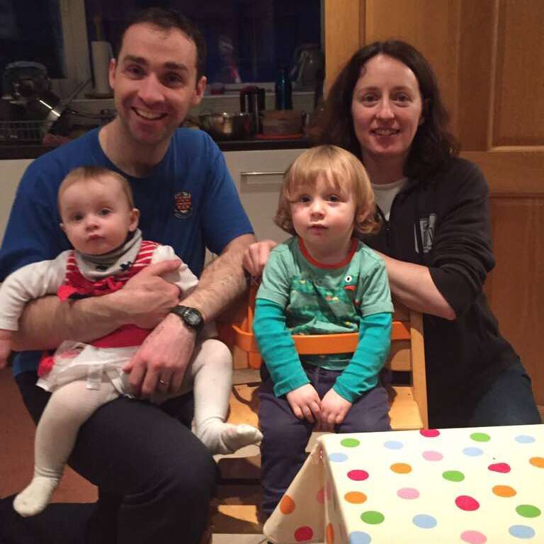 La imagen muestra a una familia de cuatro miembros posando para una foto en una cocina, con un bebé a la izquierda y un niño pequeño en una trona en el centro, rodeados de un ambiente cálido y acogedor.