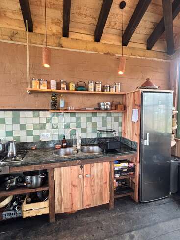 This rustic kitchen features wooden cabinets, open shelving with jars, a green-tiled backsplash, double sink, stainless refrigerator, exposed beams, and soft pendant lighting for warmth.