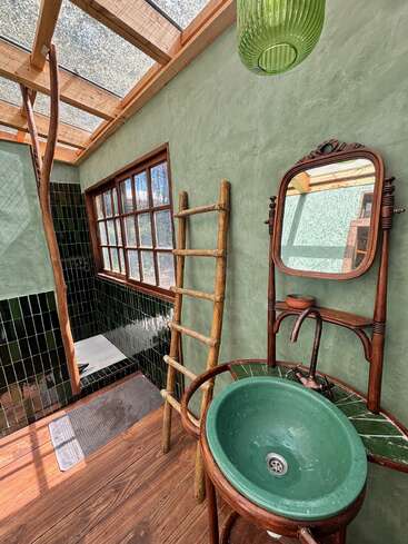This rustic bathroom features a green sink, wooden ladder, vintage mirror, natural wood elements, tiled shower, large window, and skylight, creating a cozy, nature-inspired atmosphere.