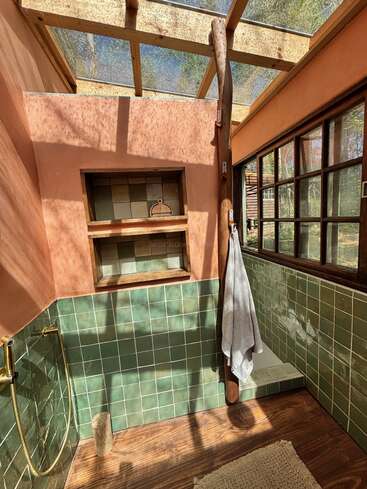 This image shows a rustic, sunlit bathroom with green tiles, wooden accents, a skylight roof, natural light, a towel hanging, and a window view outside.