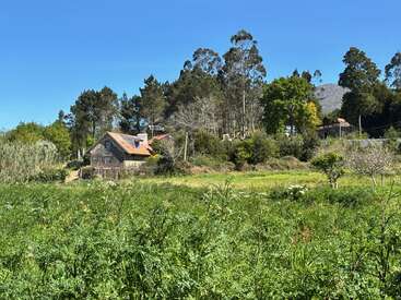 A quaint stone house with a red roof sits amidst lush greenery, surrounded by trees and fields under a clear blue sky in a peaceful countryside.