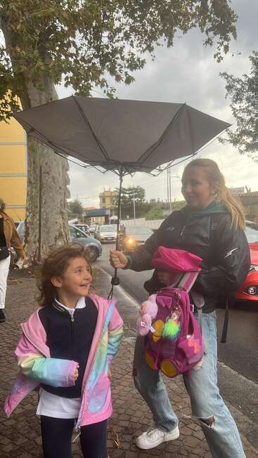 Two girls stand on a sidewalk under an inverted umbrella. One holds a pink backpack, while the younger girl smiles happily. Cars and trees are behind them.