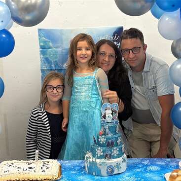 A family celebrates a girl's fifth birthday with a Frozen-themed party. Blue and silver balloons, themed cake, happy faces, and festive decorations create a joyful atmosphere.