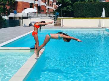 Deux filles en maillot de bain s'amusent à plonger dans une piscine bleue claire par une journée ensoleillée, entourée de bâtiments, de parasols et de haies vertes.