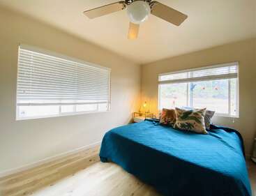 A bright, cozy bedroom with large windows, a ceiling fan, hardwood floors, a blue bedspread, and patterned pillows. Minimalist decor and a outside view.