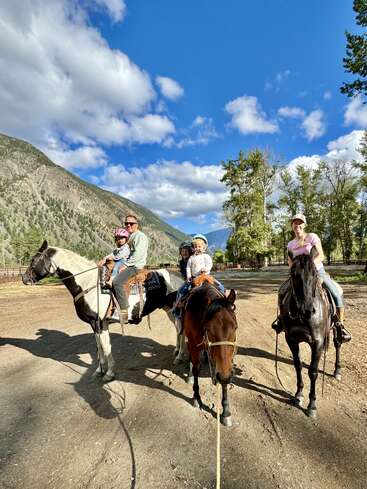 Une famille de quatre personnes, dont deux enfants, monte à cheval sous un ciel bleu lumineux avec des nuages épars, entourée de montagnes et de grands arbres, pour profiter de la nature.