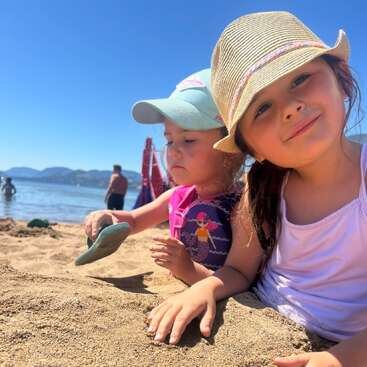Deux jeunes filles jouent joyeusement dans le sable sur une plage ensoleillée, portant des chapeaux pour se protéger du soleil. Le ciel et l'eau d'un bleu éclatant forment une magnifique toile de fond.
