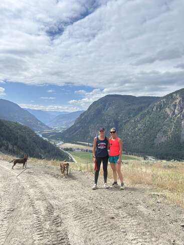 Deux femmes se tiennent sur un chemin de terre surplombant une vallée montagneuse pittoresque, accompagnées de deux chiens, sous un ciel partiellement nuageux et entourées de collines verdoyantes.