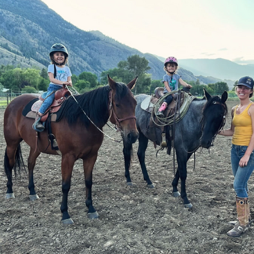 Deux jeunes filles portant des casques montent joyeusement à cheval, tandis qu'une femme souriante vêtue d'un débardeur jaune se tient à côté d'elles, tenant un cheval à l'extérieur. Montagnes en arrière-plan.